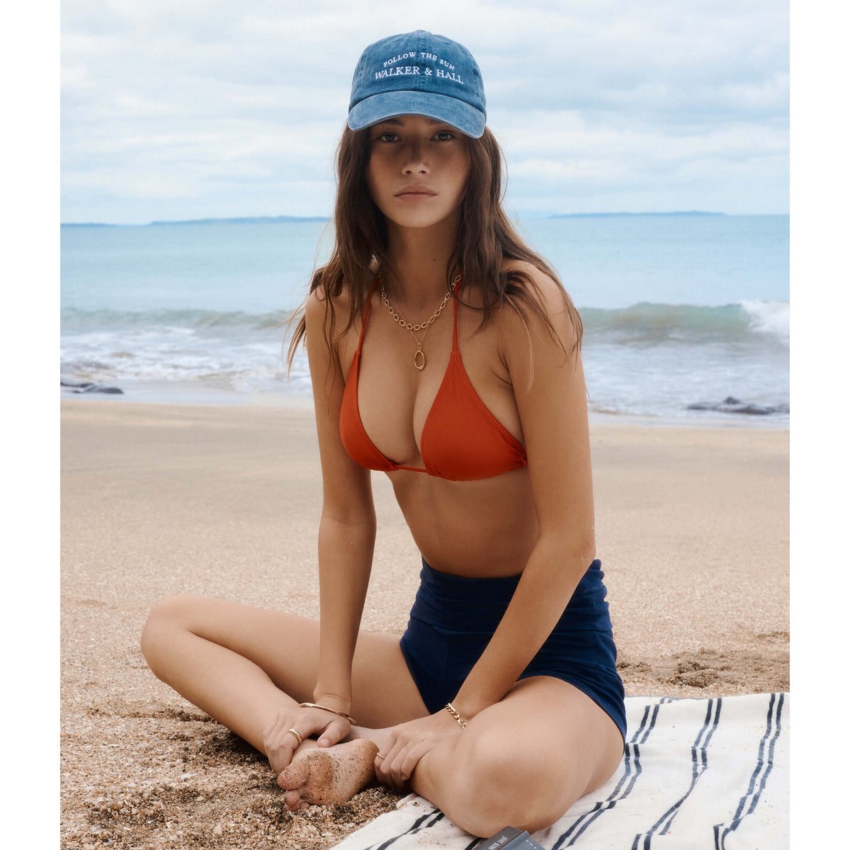 Model sitting on NZ beach wearing Walker & Hall Follow The Sun cap and gold jewellery