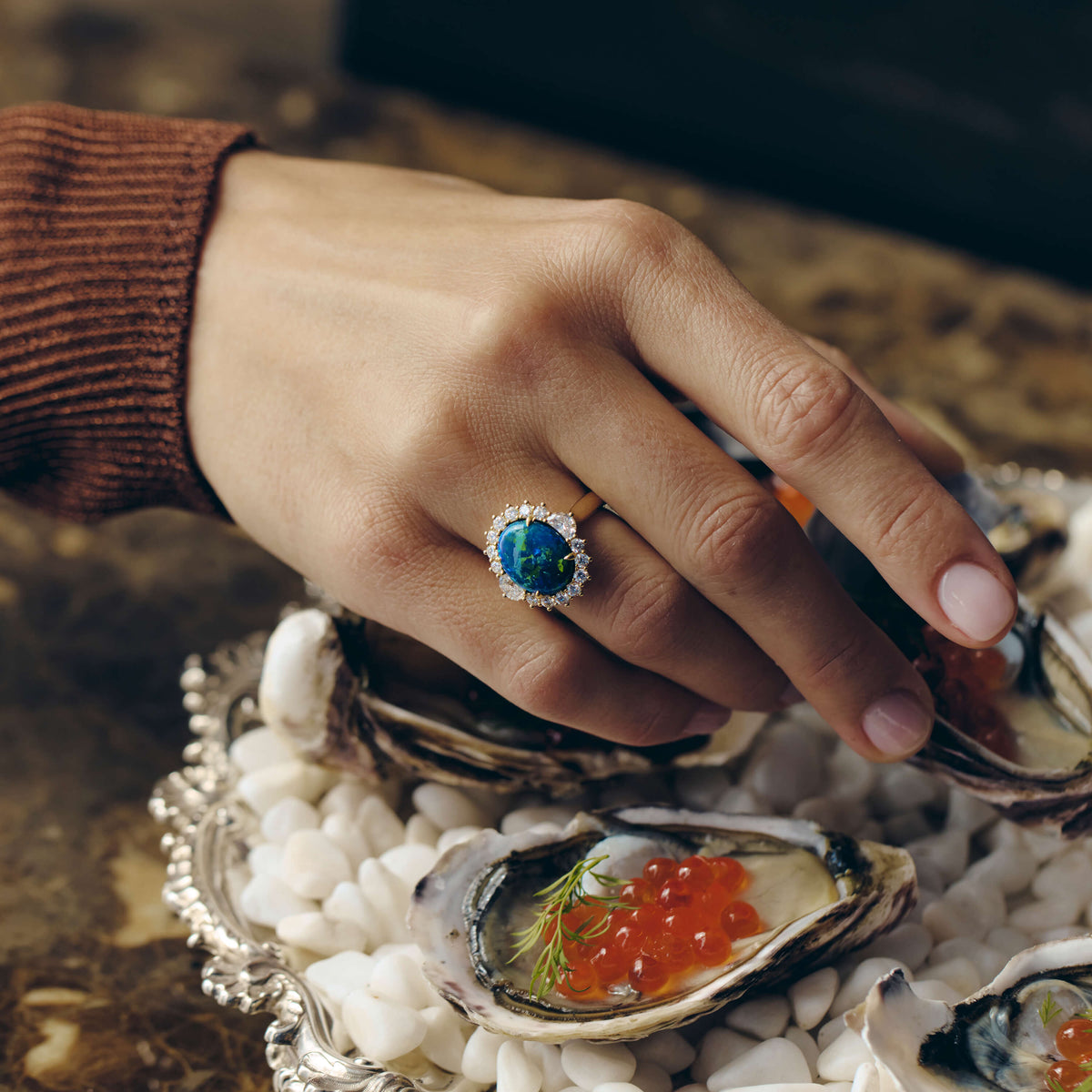 Hand wearing an opal and diamond ring holding an oyster with caviar on a marble table